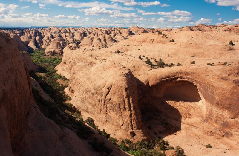 The image shows a vast, arid landscape with sandstone formations. The rock formations are rounded and sculpted by erosion, creating a unique and rugged terrain. A natural arch is visible in the foreground, adding to the geological interest. Patches of green vegetation are sparsely distributed in the valleys and crevices, providing a contrast to the dominant reddish-brown hues of the rocks. The sky is blue with scattered clouds.